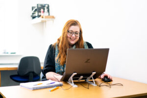 A girl sits at a laptop on a computer desk. She has long red hair.