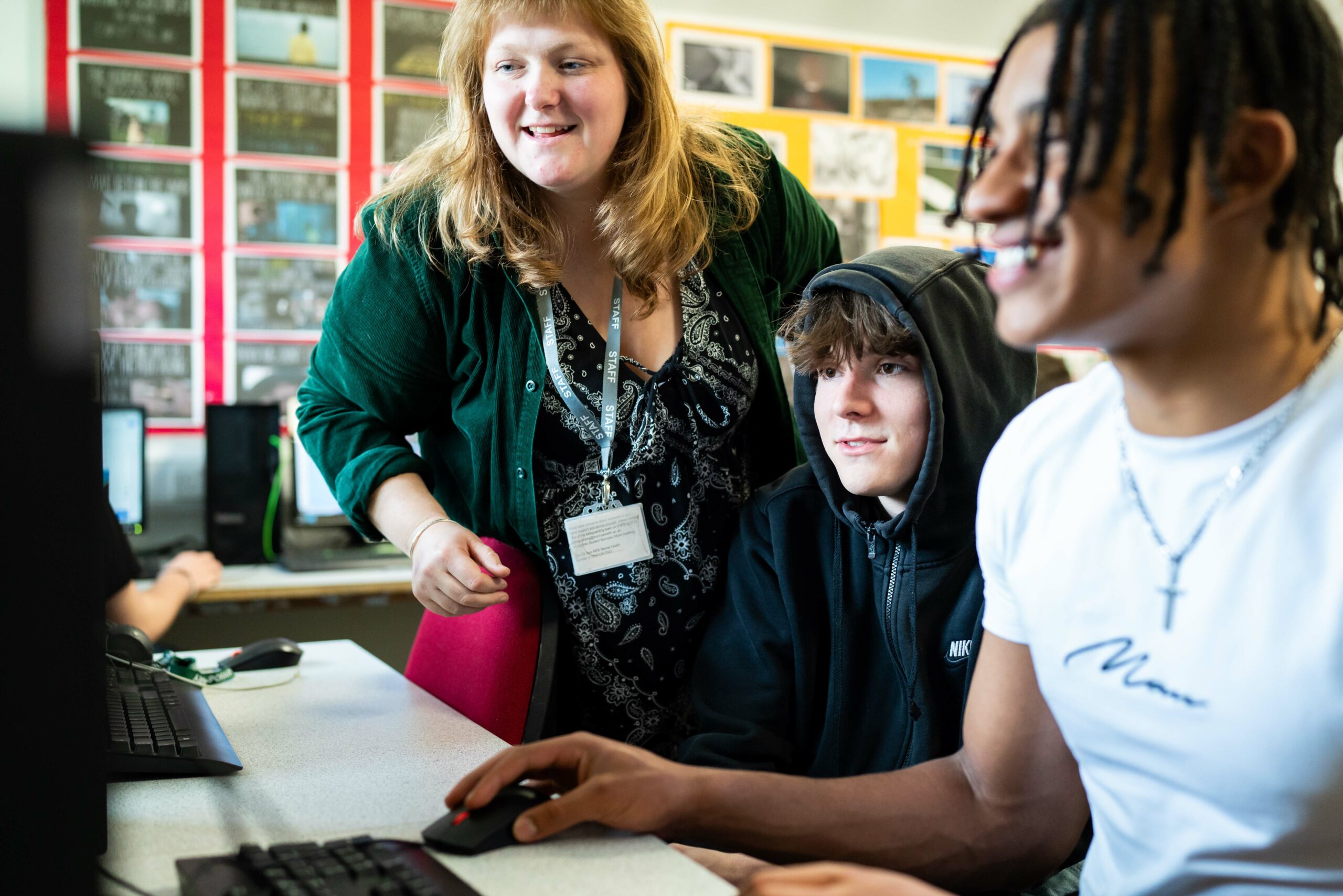 A student smiles while working at his computer. Another student and a lecturer are sat next to and behind him, also looking at the first student's computer screen which we cannot see as the viewer.