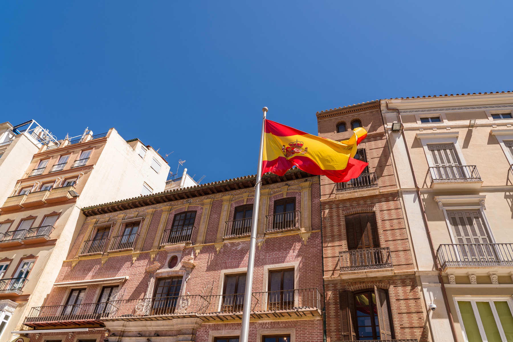 Spanish flag in the old town of the city of Malaga. Beautiful typically Spanish buildings architecture in background. Blue sky in a sunny summer day