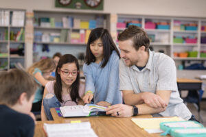 A male teacher sits a desk and helps two of his students with their school work. There are others working in the background.