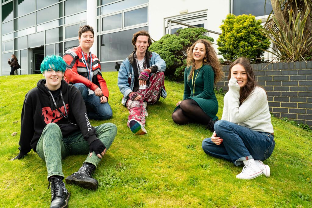 A group of students sit on the grass facing the camera. They all smile.