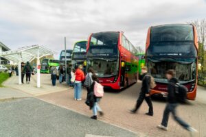 A line of buses wait at the Truro campus bus bay while students walk by. The students are blurred with motion.