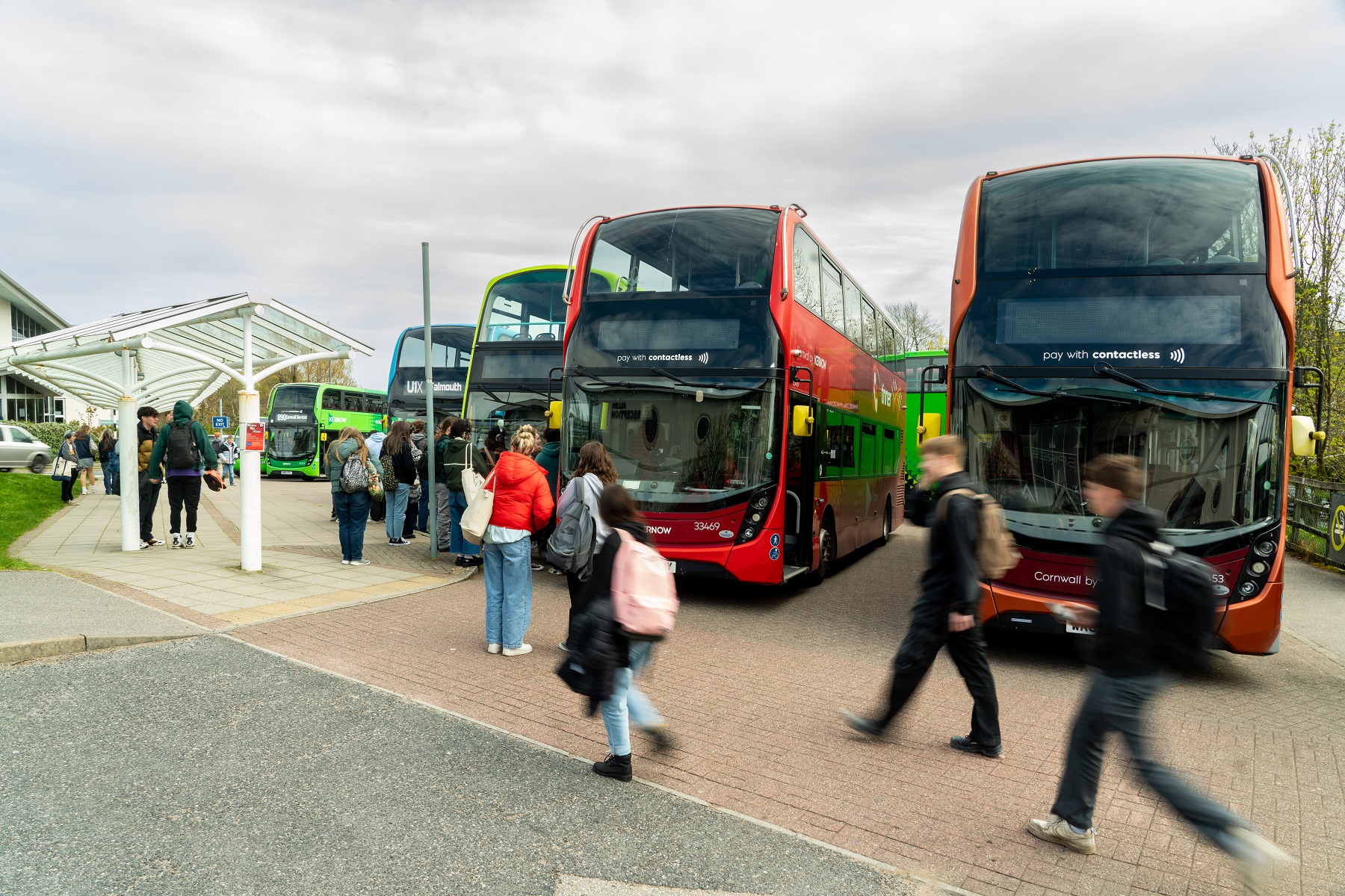 A line of buses wait at the Truro campus bus bay while students walk by. The students are blurred with motion.