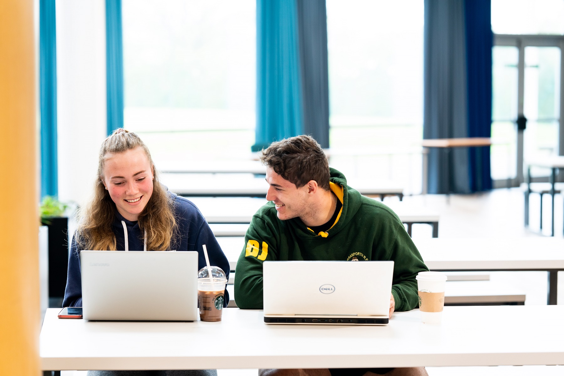 Two students sit at a table with their laptops and cold drinks in the Fal building canteen at the University Centre Truro and Penwith.