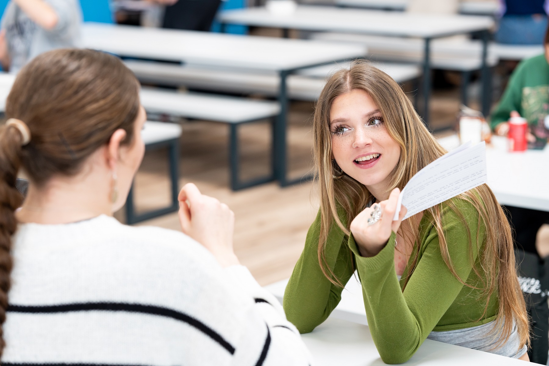 Two smiling students chat together at a table. One student holds a piece of paper in her hand.