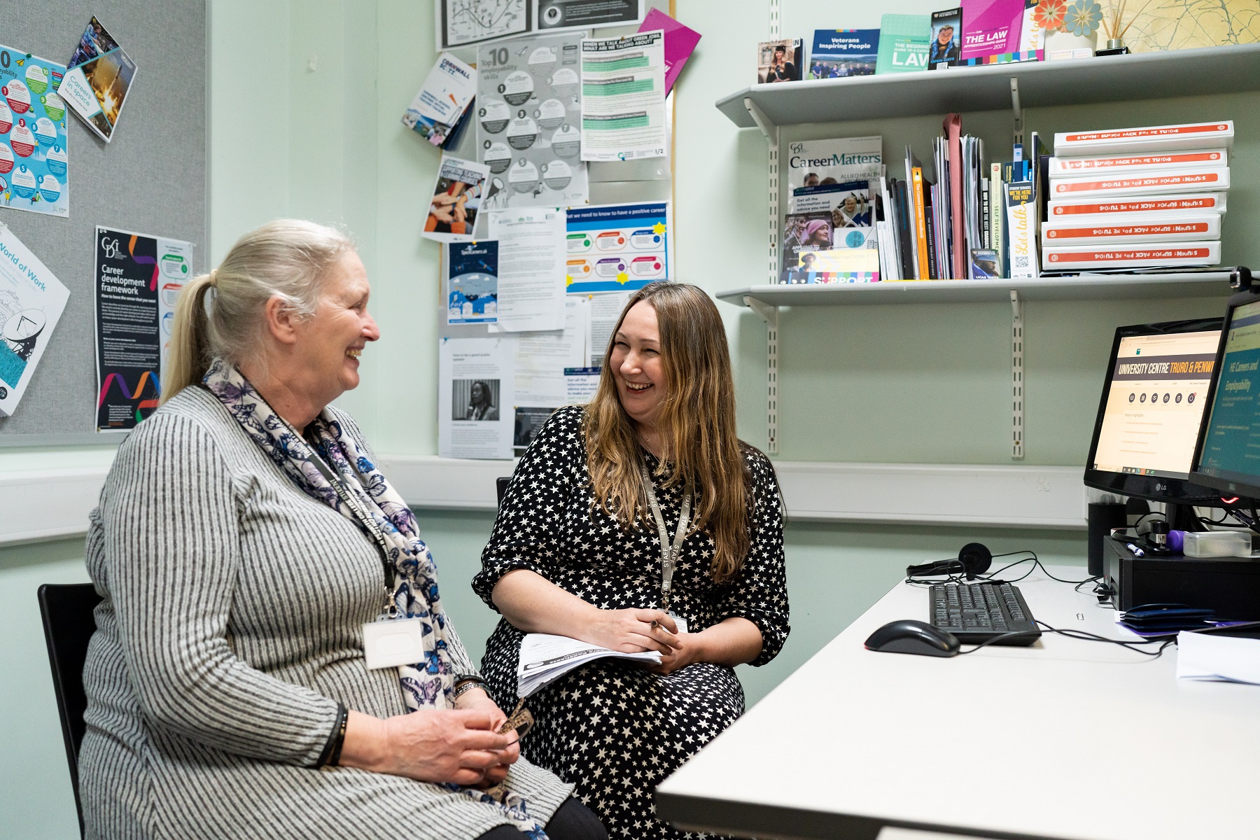 An adult University student and a member of staff chat and laugh in an office.