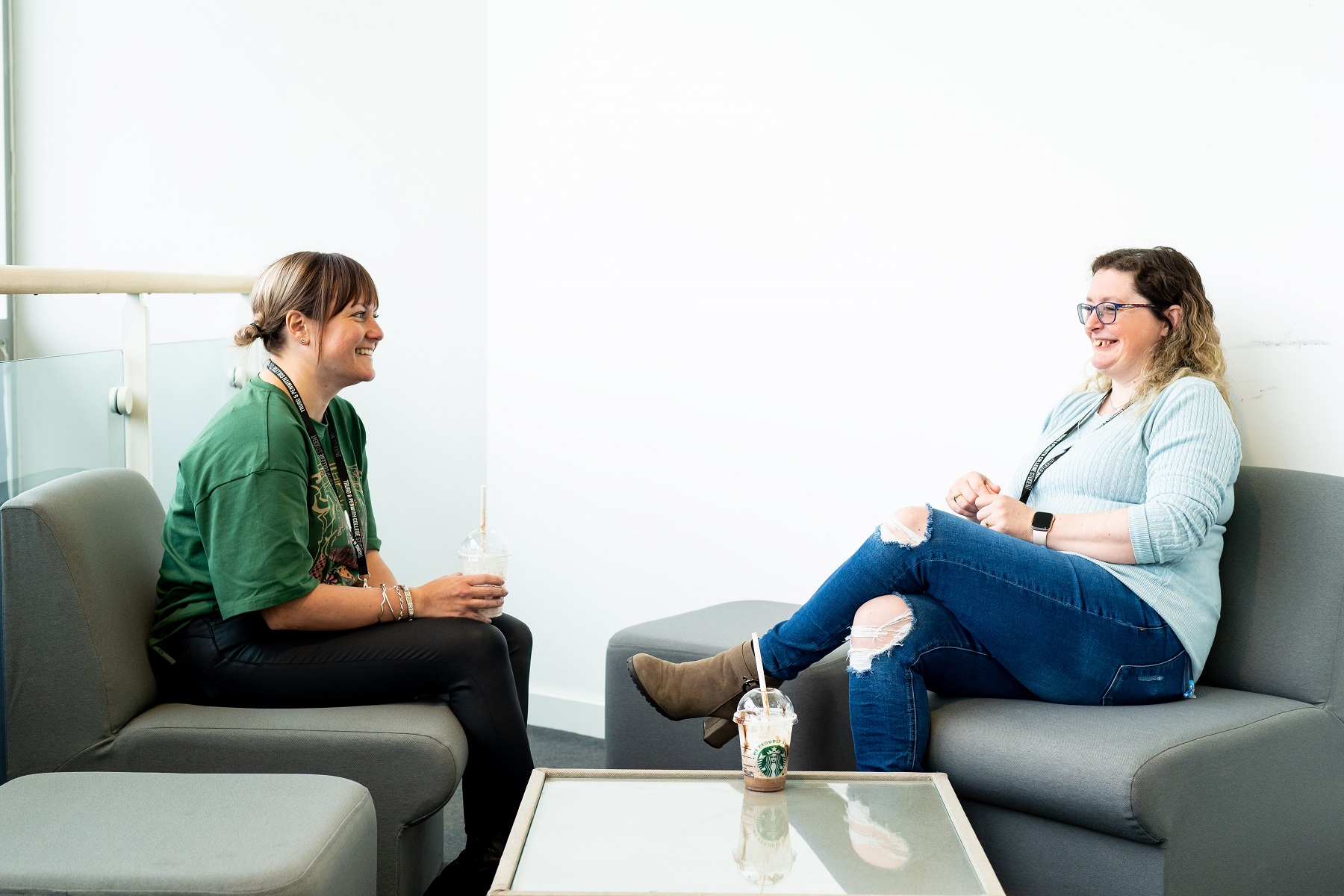 Two smiling adult University students sit in comfortable seating with a drink each.