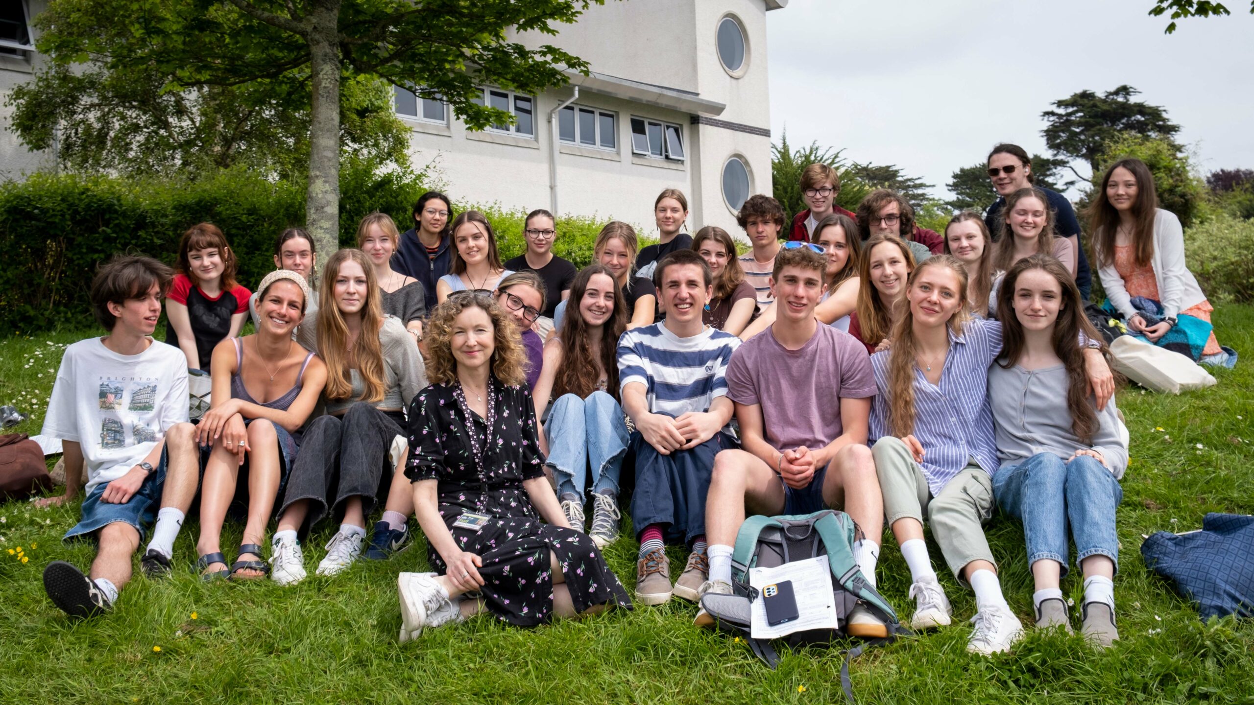 The IB cohort from Truro College smile for a picture while sitting outside on a grassy bank.