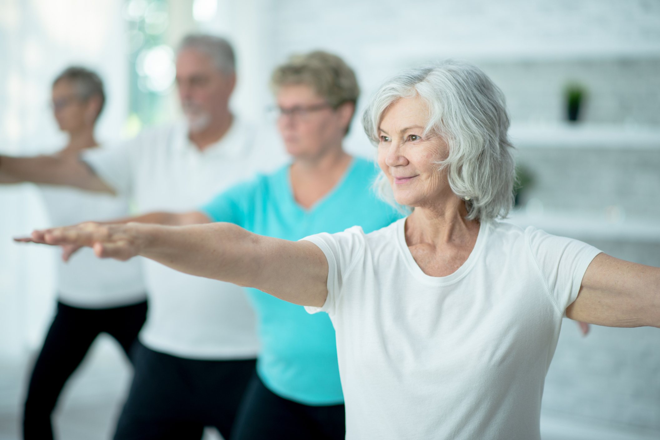 A small group of seniors stand evenly spready apart in a studio with their hands extended as they stretch and balance during a Tai Chi class.  They are each dressed comfortably and are focused on the movements.