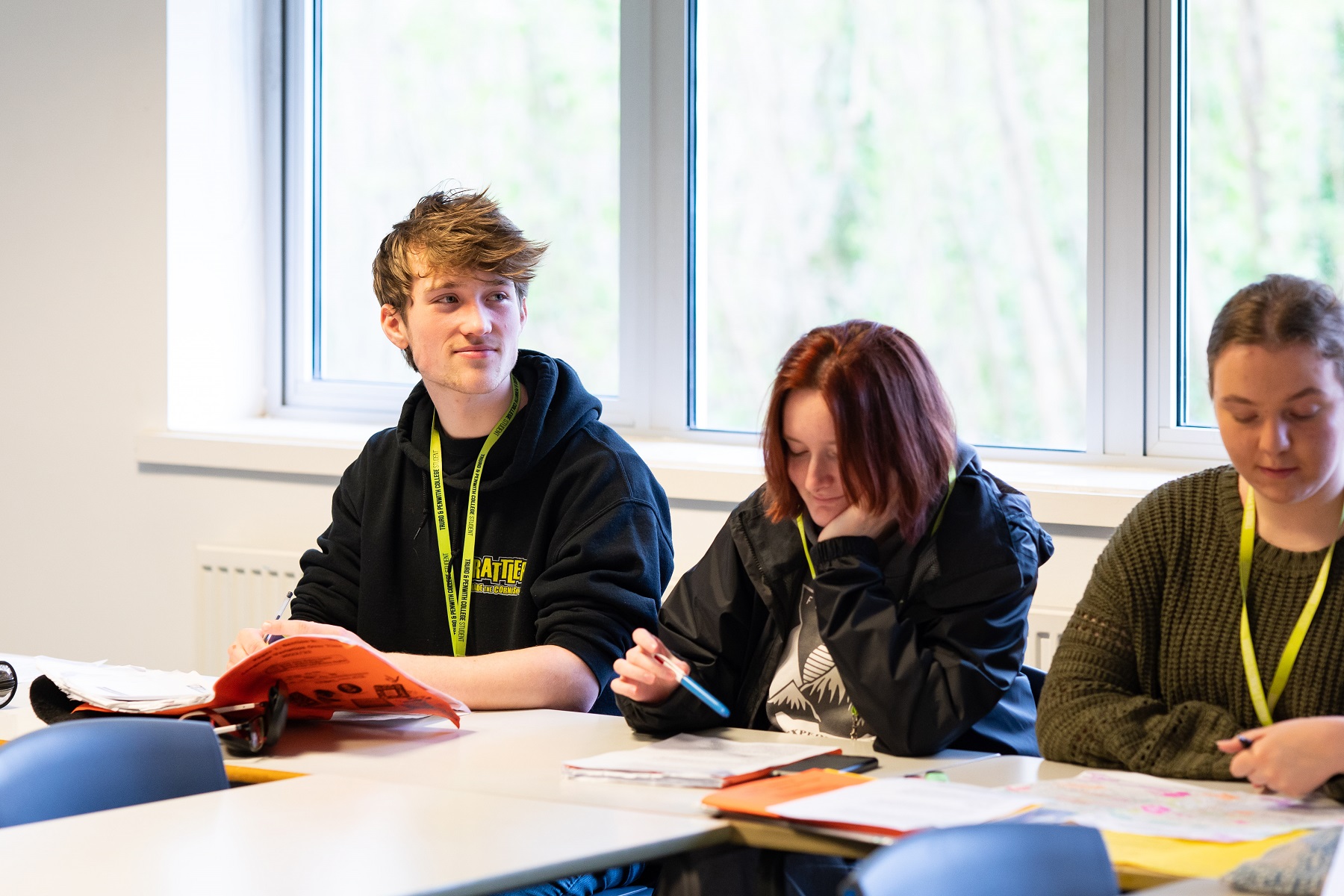 Three students sat working at a classroom table with books and notepads in front of them. They are all wearing bright student lanyards.