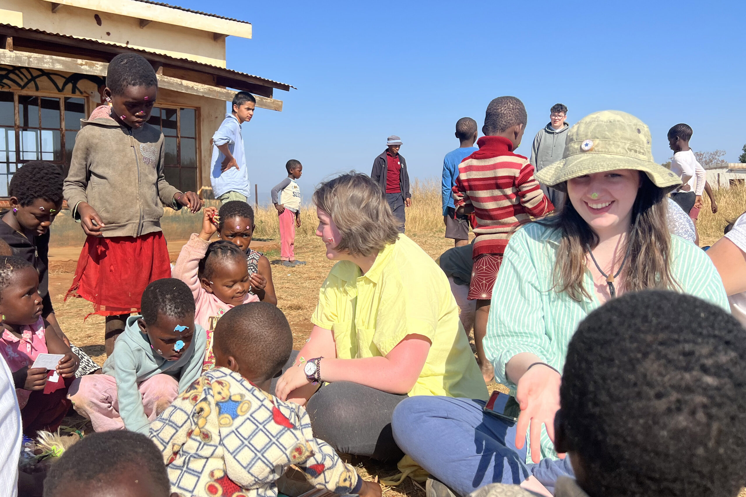 Students sit outside on the grass with children from Eswatini making crafts and playing with stickers.