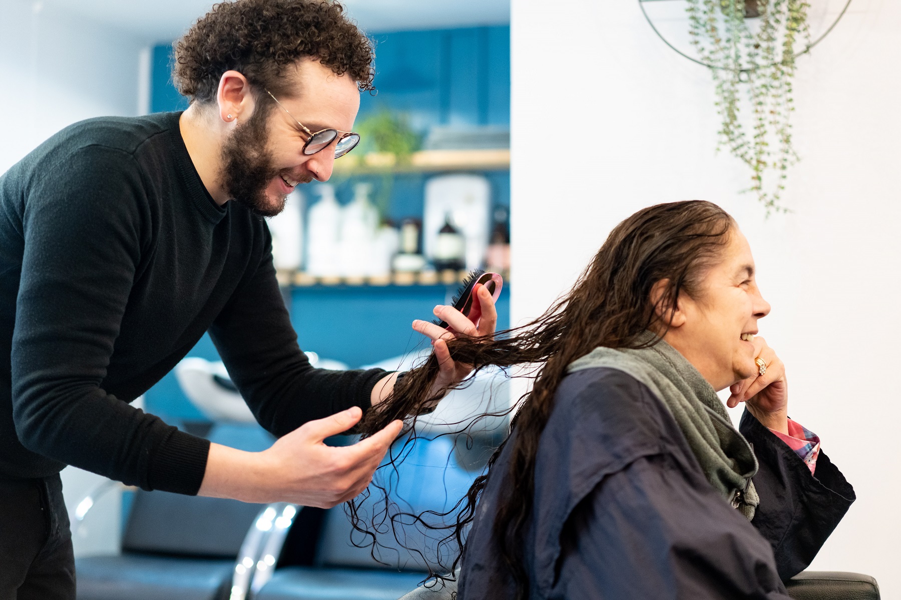 A male hairdresser combs out a woman's long, wet hair with a hairbrush within a salon setting.