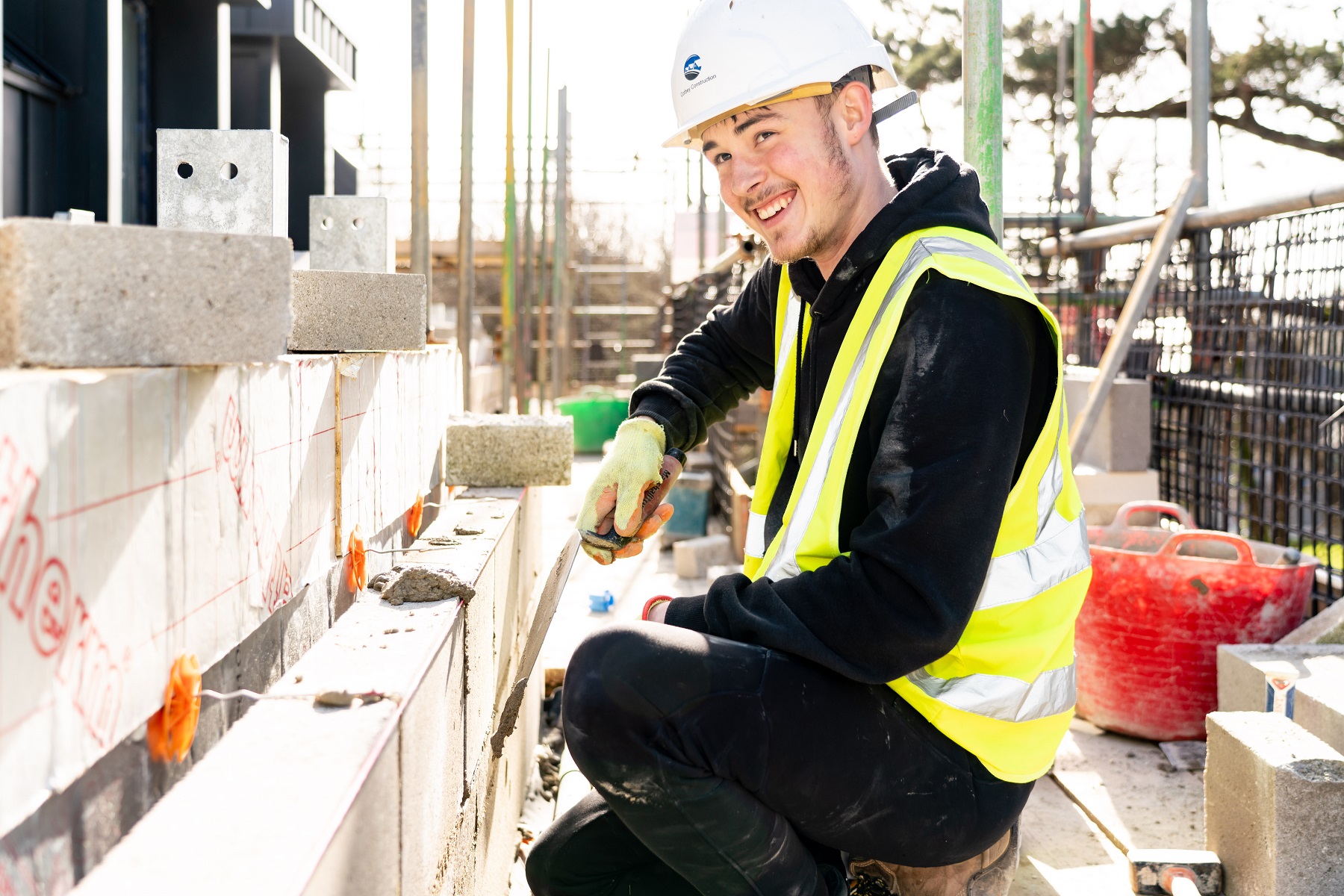 A young man in a high vis vest and hard hat lays bricks in a construction setting.