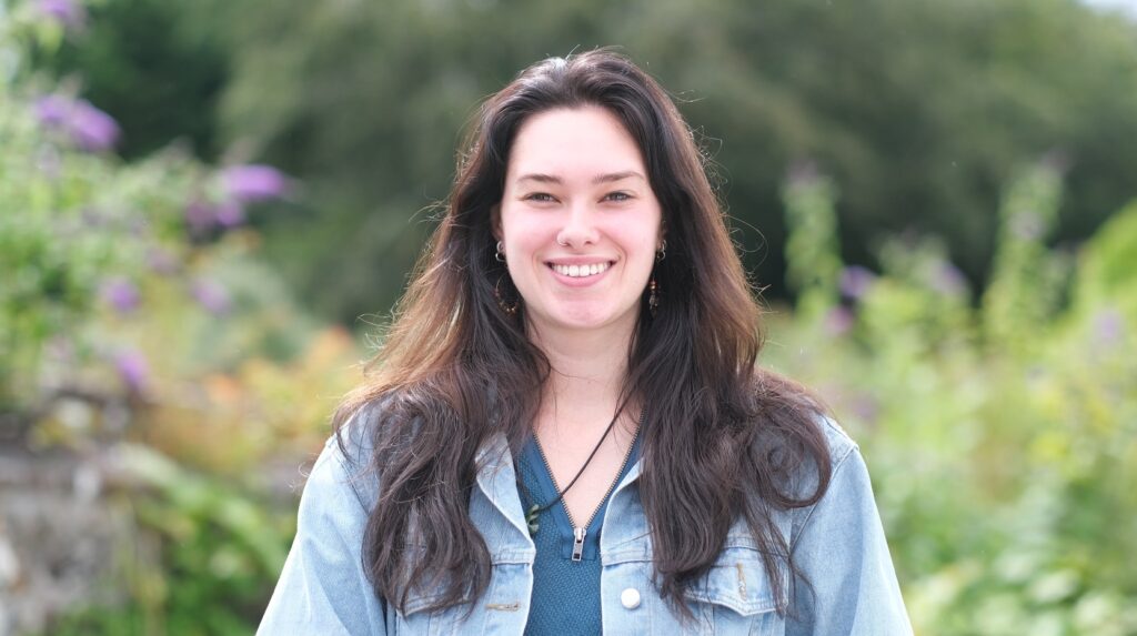 A young woman with long dark brown hair wearing a blue top and pale blue denim jacket smiles outside with green and pink floral bushes behind her.