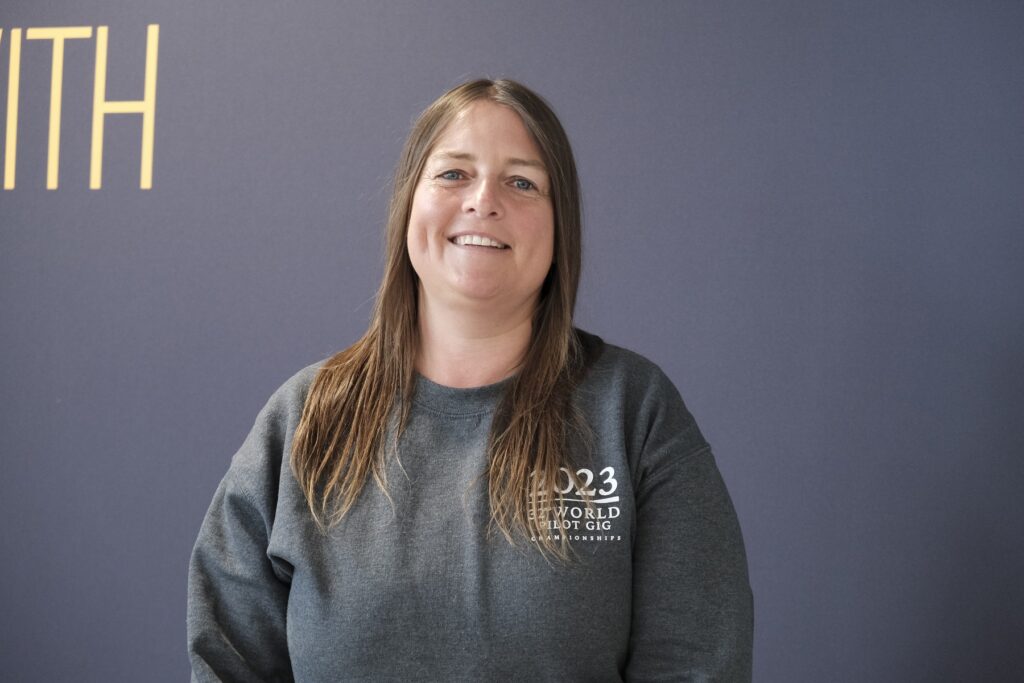 Kat BA Hons Education & Training. A student with brown shoulder length hair wearing a grey jumper smiles looking at the camera. She is stading infront of a black banner.