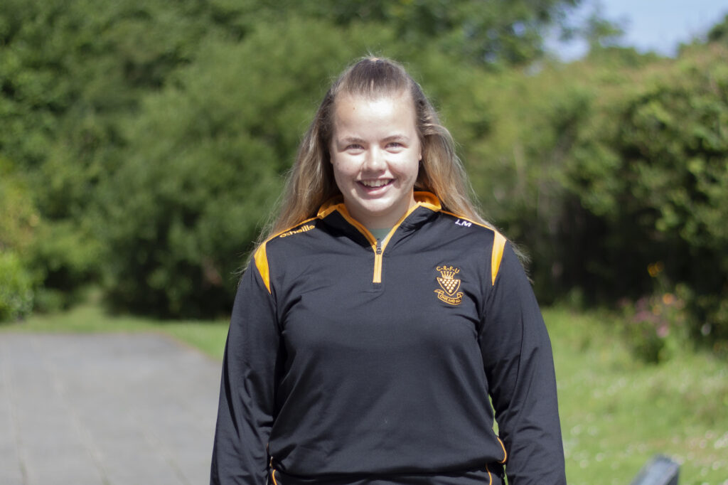 Lucie looks at the camera smiling on a bright sunny day. She is wearing a black rugby shell jacket.