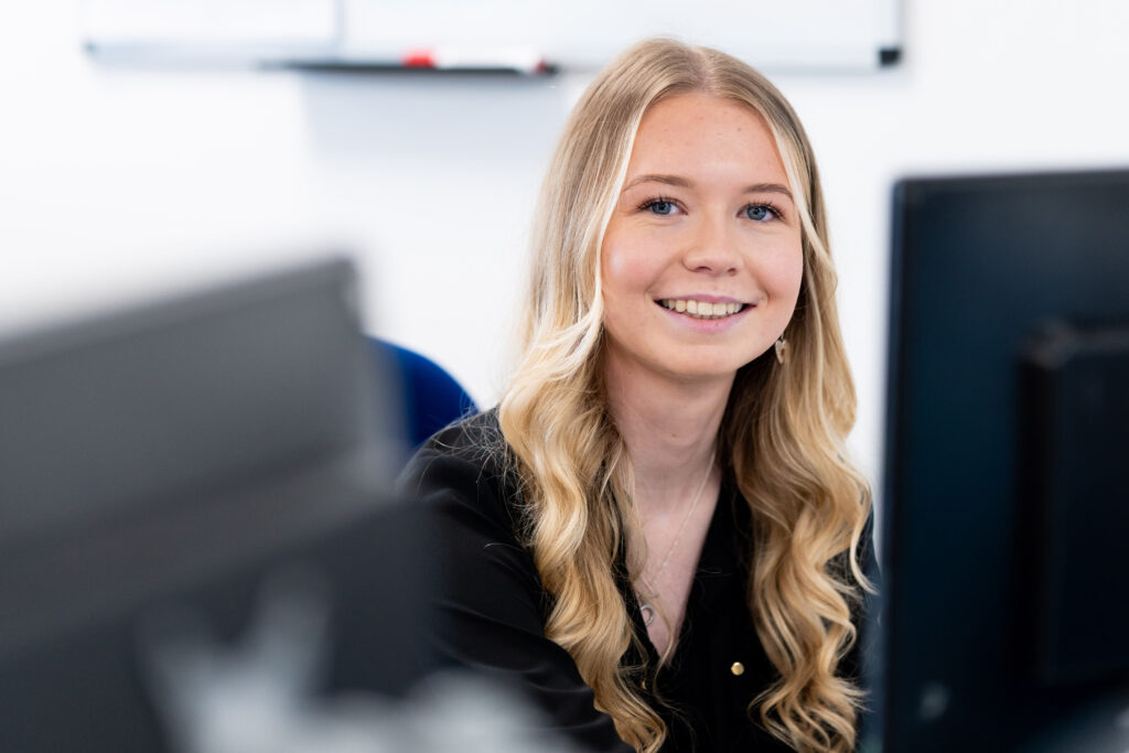 A picture of Olivia at her desk in front of a computer looking at the camera and smiling.