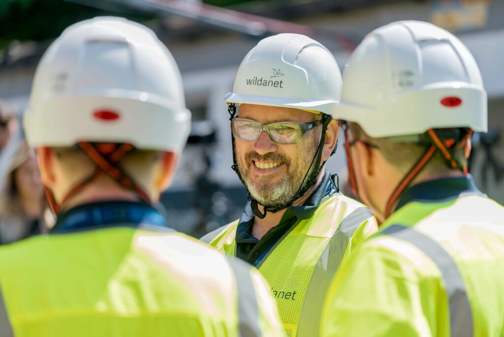 Telecoms Field Operatives in conversation, wearing yellow hi-vis, white hard hats and safety glasses.