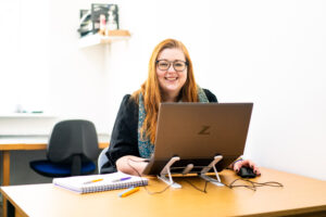 A female with glasses sits at a silver laptop at a desk and smiles down camera.