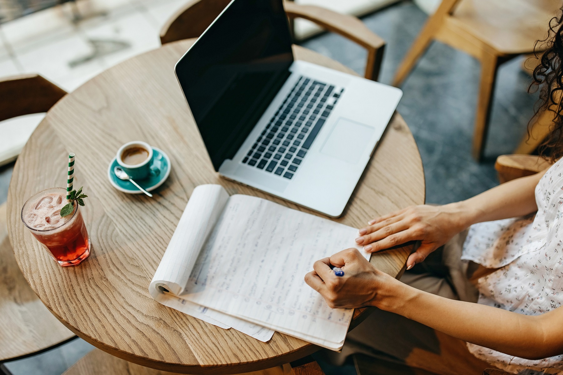 Young woman siting at caffe, busy writing in notebook, studying and finishing work. There is laptop, juice and coffee on the table.