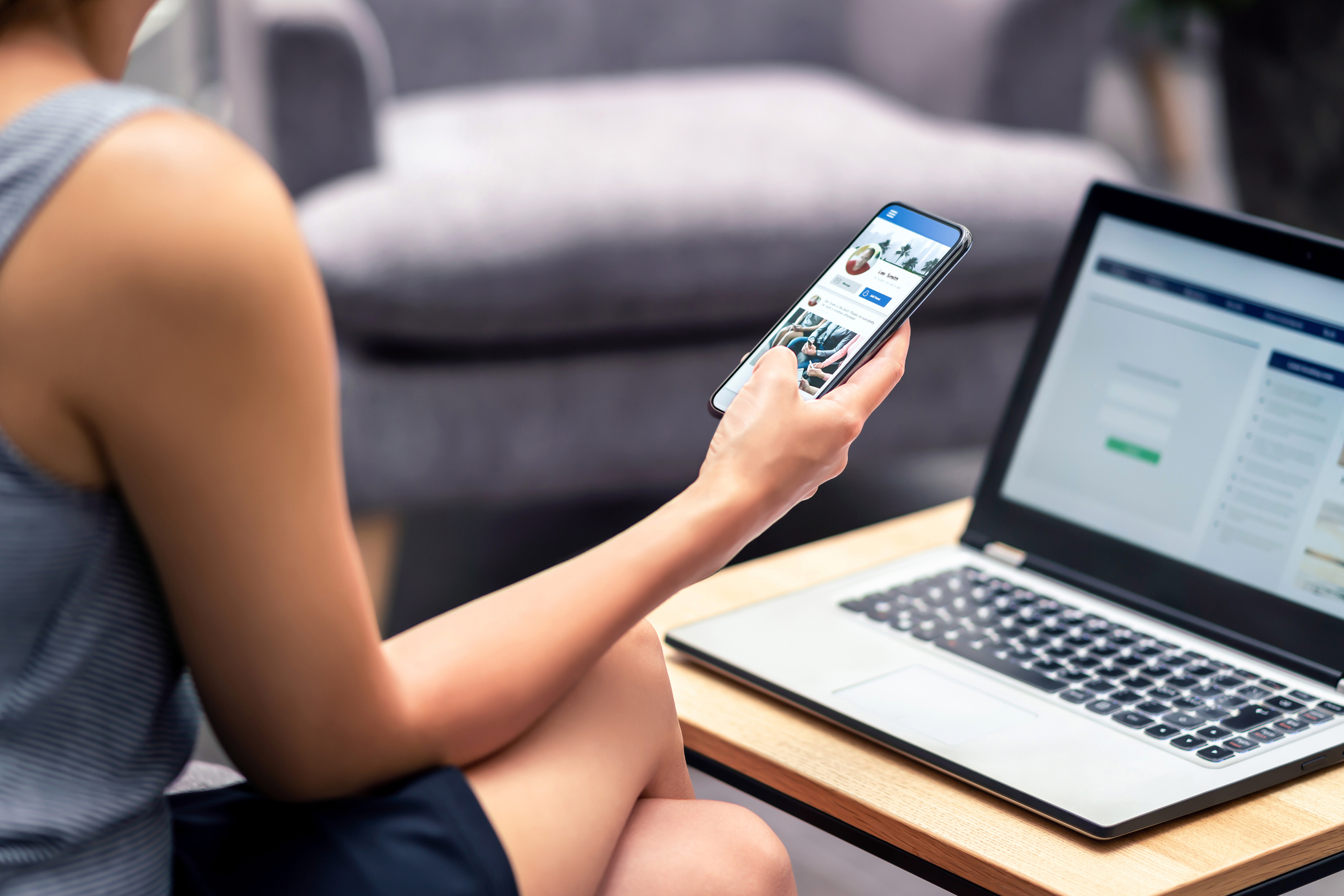 Woman looking at social media feed on mobile phone with laptop open in front of her on a coffee table.