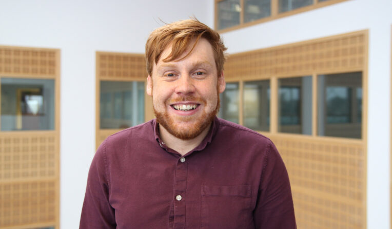 A Student with short red hair wearing a purple shirt smiles at the camera