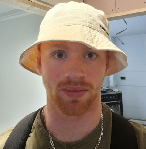 A student with short beard wearing a beige bucket hat and a green t-shirt looks at the camera.