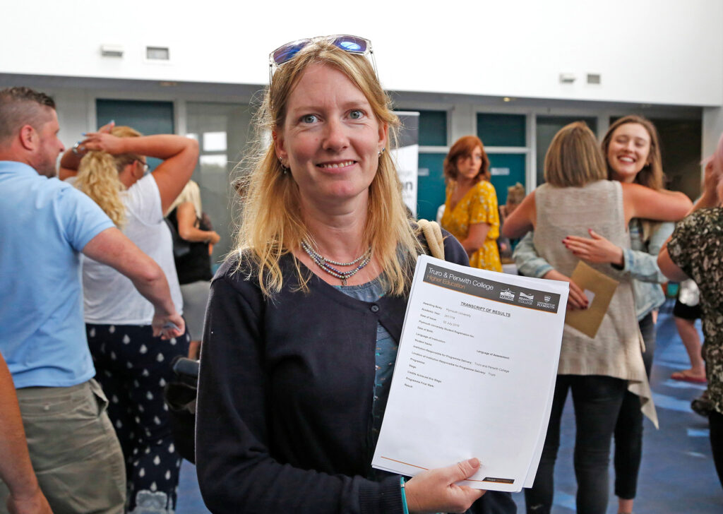 A sudent with shoulder length blonde hair holds her results up to the camera and smiles