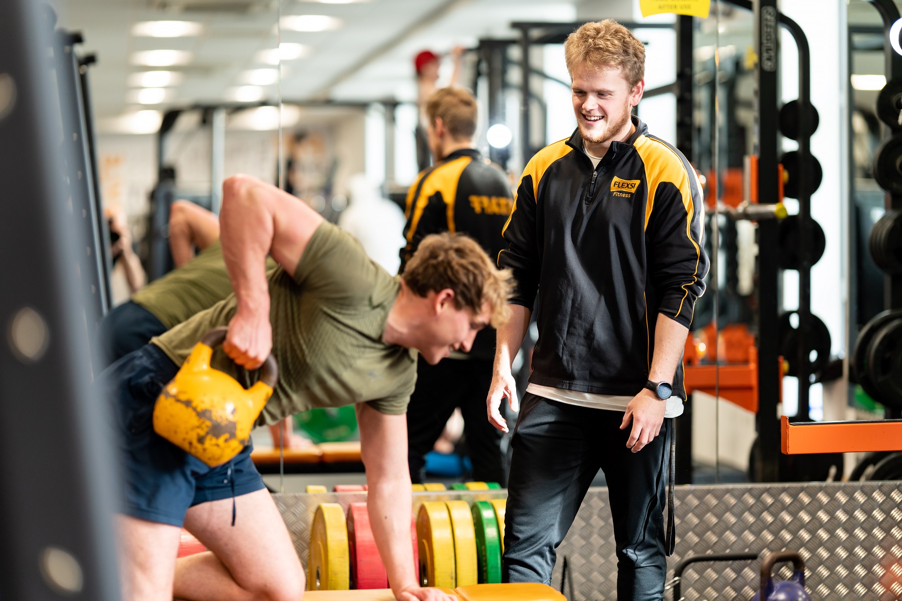 An apprentice stands in a gym mentoring a customer who is lifting a kettlebell.