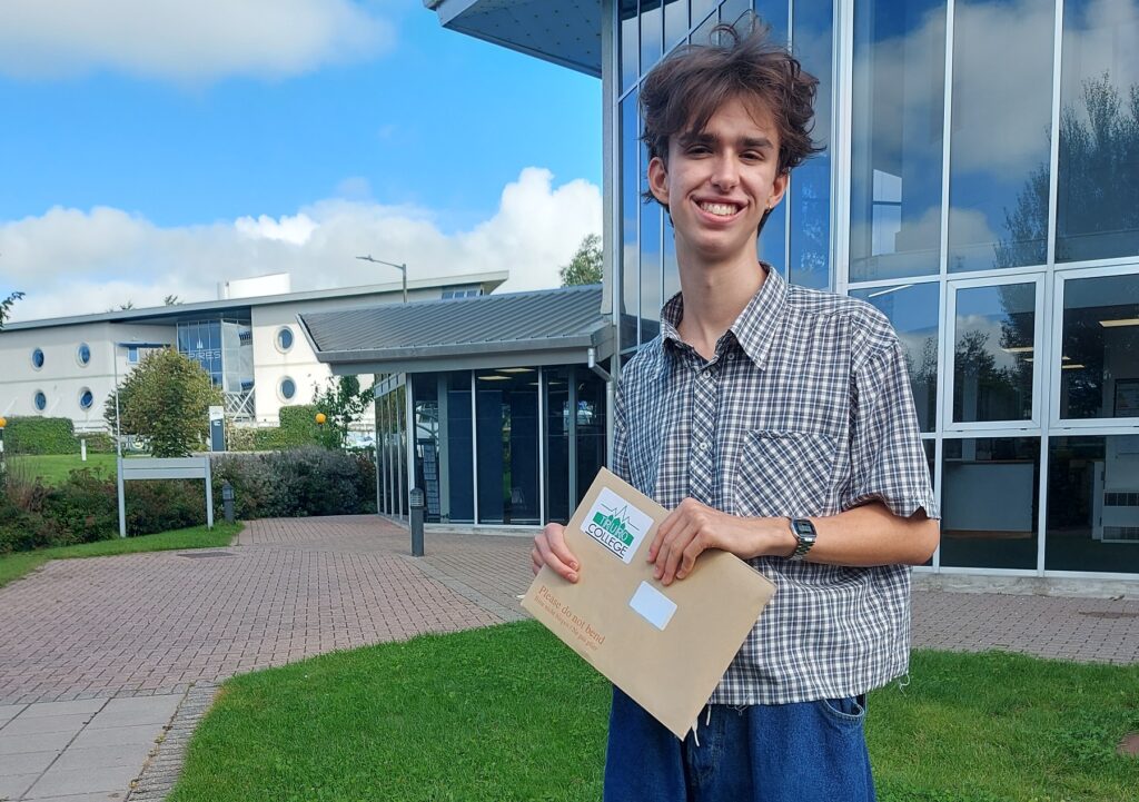 Gabe smiles down camera with the Kenwyn and Allen buildings in the background. He is wearing a checked shirt, blue jeans, holding his results envelope.