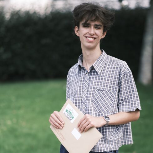 Student Gabe, smiles down camera, standing in a green area of the campus with a checked short sleeved shirt and his results envelope in hand