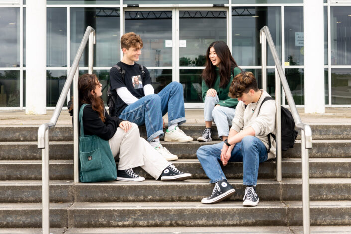 A group of students sit on the steps of one of the college buildings, laughing and chatting.