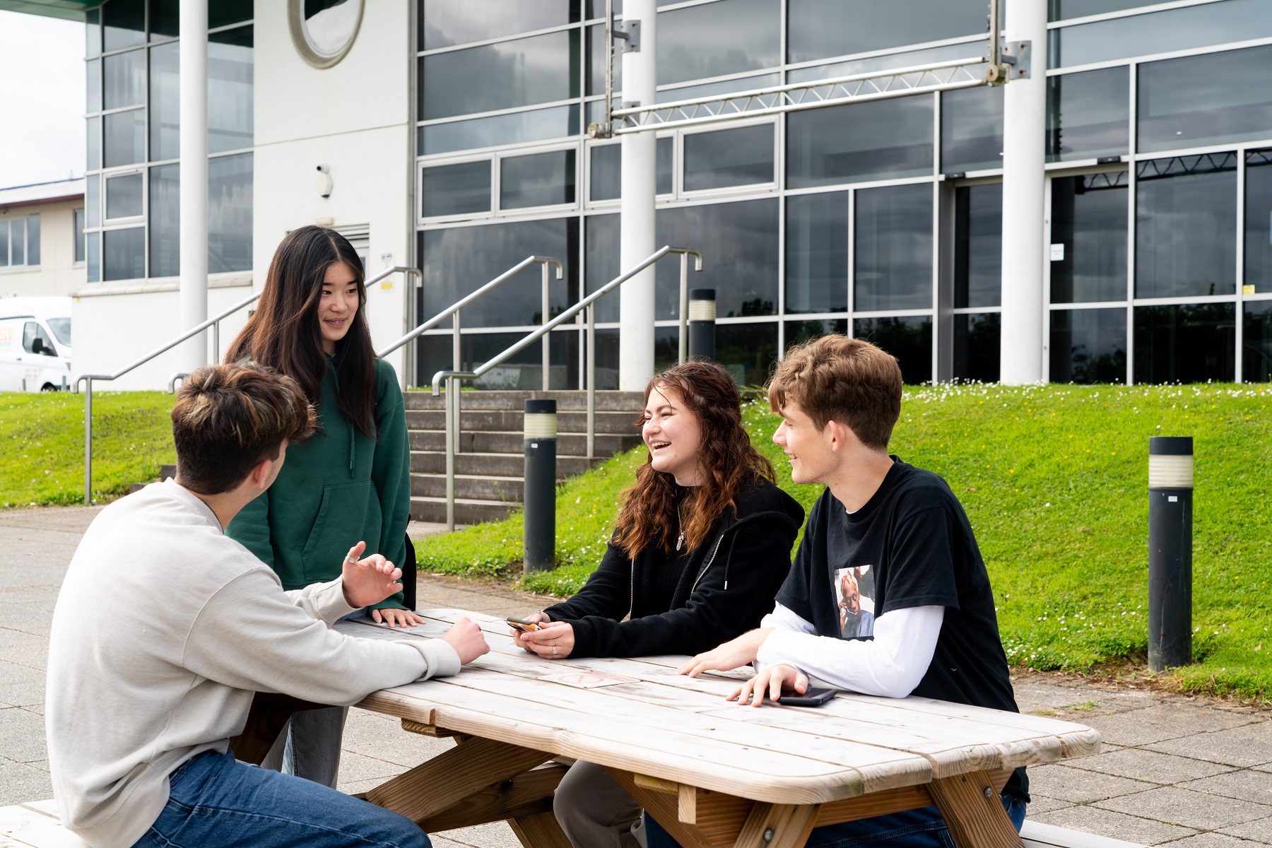 A group of students sit at a picnice bench outside a Truro campus building chatting and laughing.