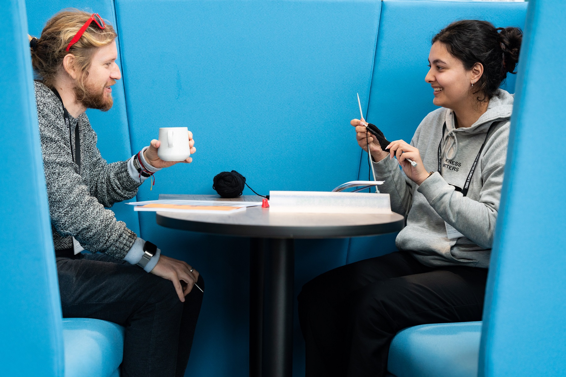 Two students sit in a seating booth at the University Centre Truro and Penwith. One has a mug in their hands while the other is knitting. Both smile at each other.