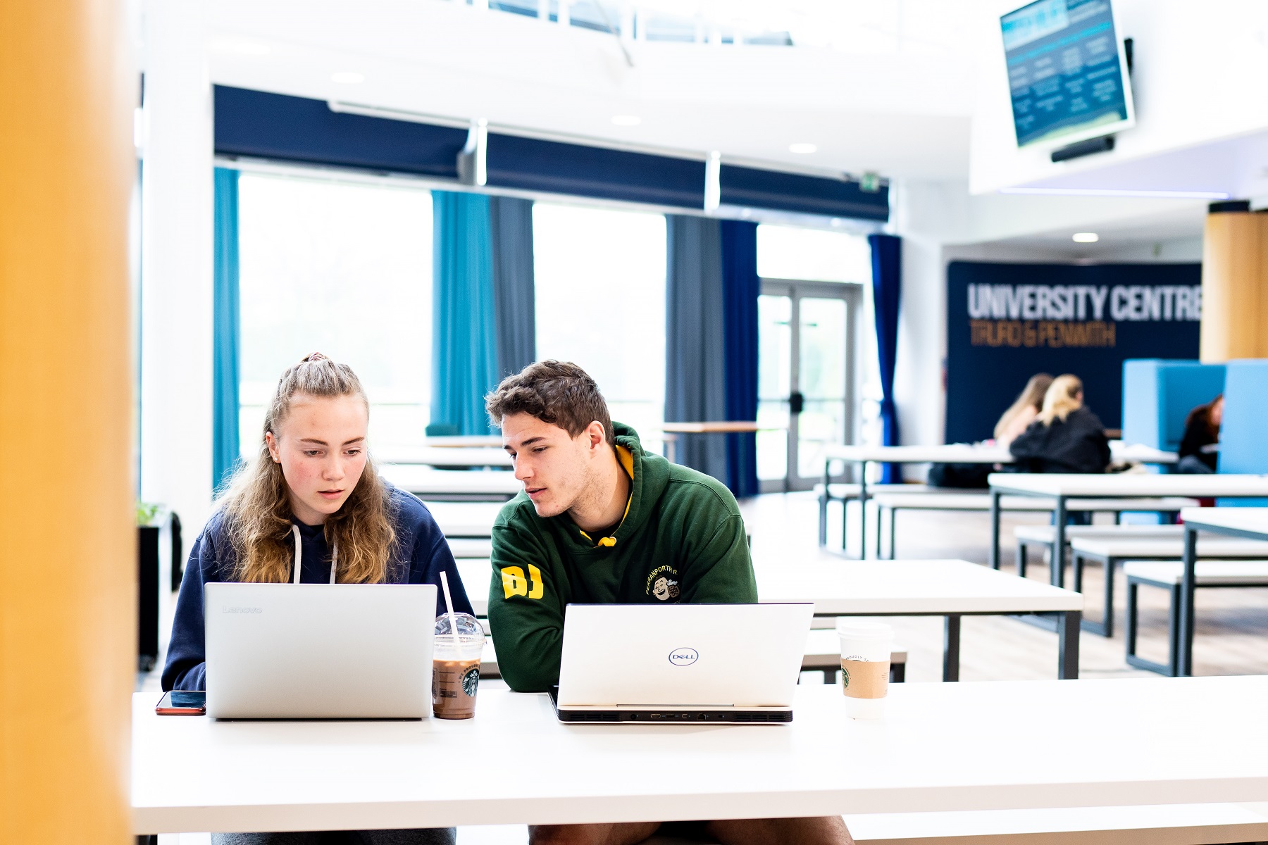 Two Higher Education students work on laptops in the canteen area of the Fal building.