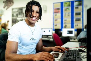 A boy looks at the camera and smiles while he uses a computer in front of him.