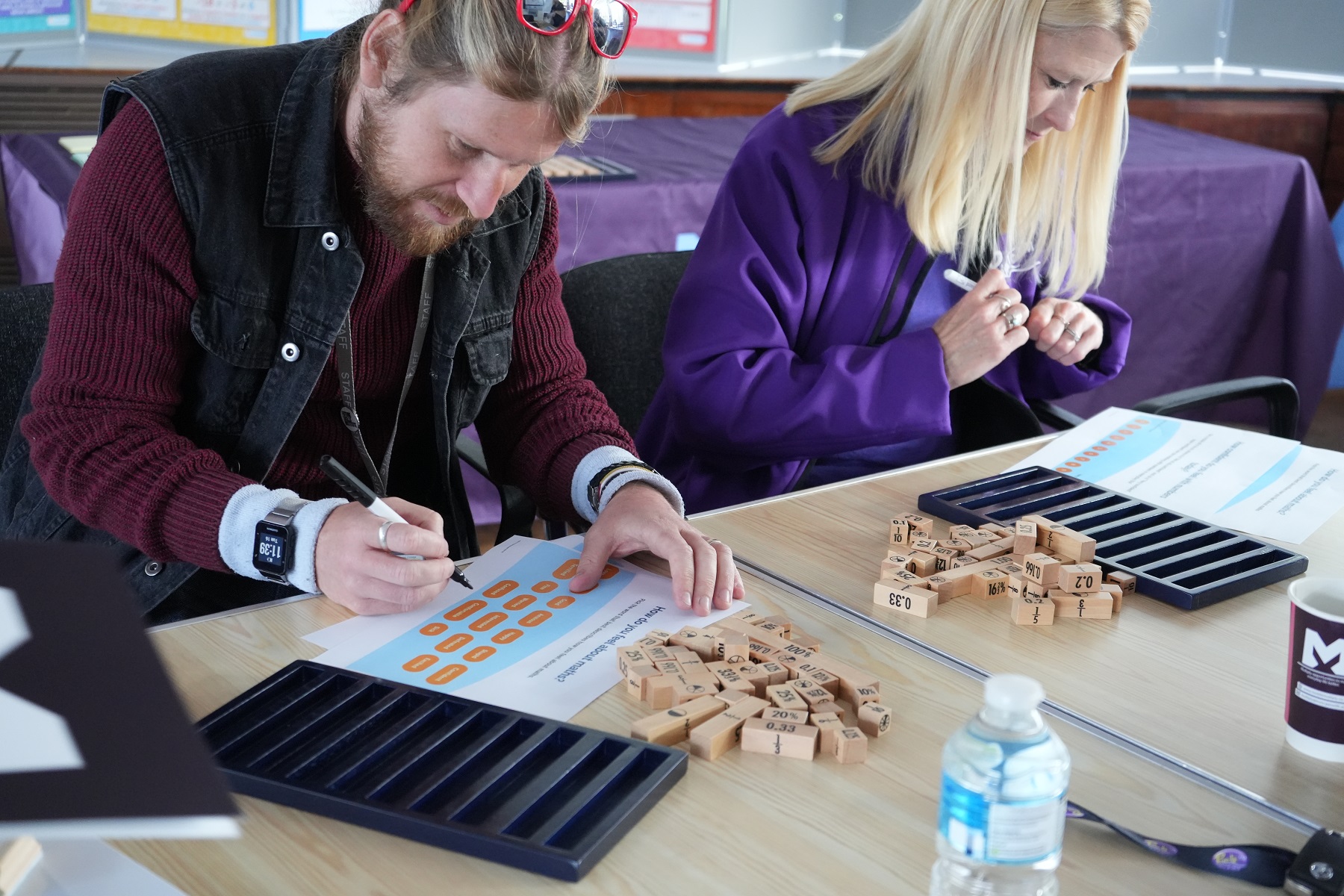 Two adult students sit at a classroom table working out mathematics problems as part of the Multiply programme. There is an assortment of maths equipment on the table.