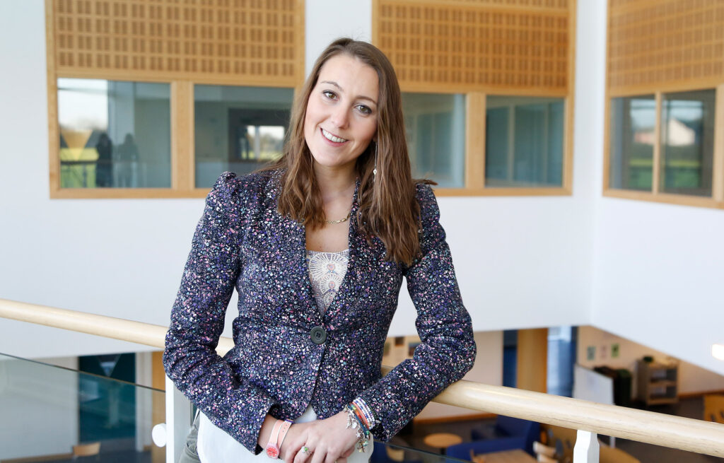 A University student wearing a blue jacket with shoulder length brown hair smiles at the camera