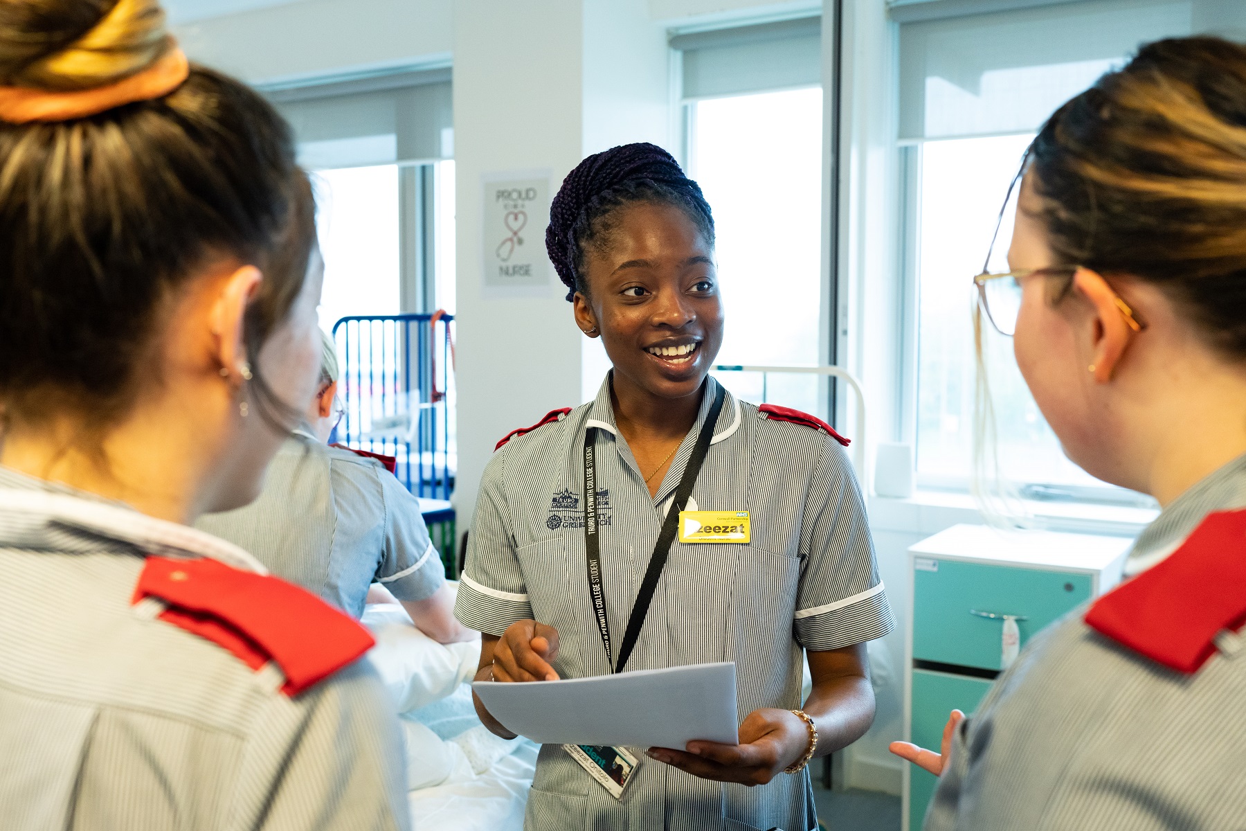 A student in a nurse's uniform branded with the University Centre Truro and Penwith and University of Greenwich look to fellow students with a paper in her hands.