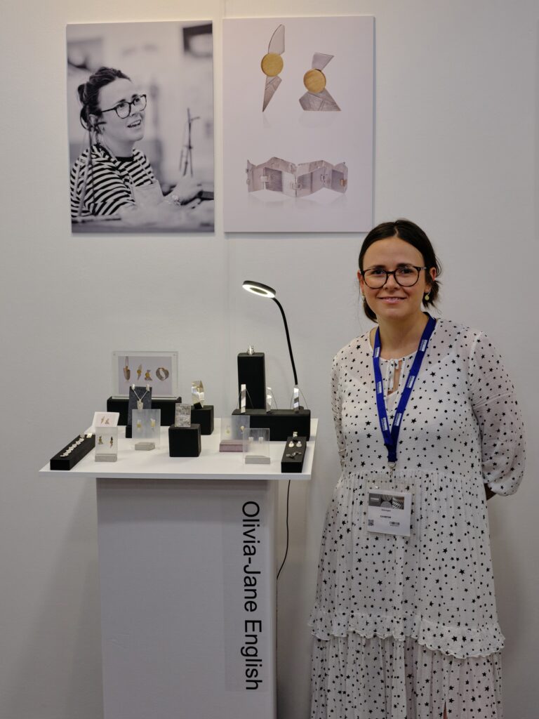 A studentwith dark hair wearing a long white dress with black stars wearing glasses poses next to a stand of handmade jewellery at an exhbition