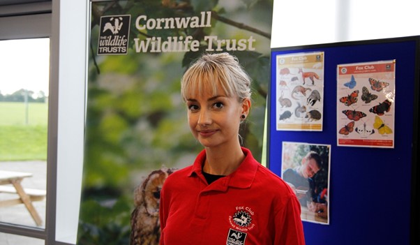 A young woman with blonde hair tied behind her hed wearing a red polo top smiles at the camera infront of a Cornwall Wildlife Trust banner