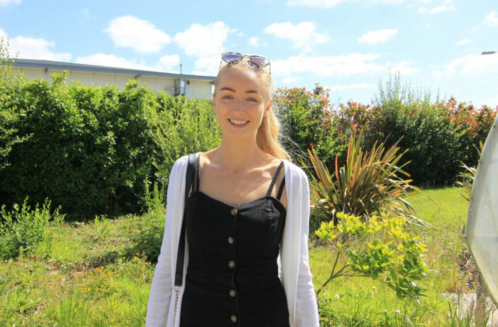 A student with blonde hair in a ponytailwearing a black denim dress with a white cardigan smiles at the camera. There are bright green bushes as the background.