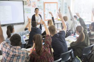 Rear View Of Teenage Students Raising Hands In Classroom