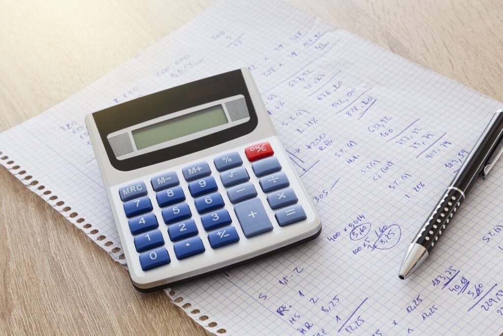 On a wooden table is a sheet of notebook squares with handwritten numbers and calculations and on the sheet is a pen and a calculator.