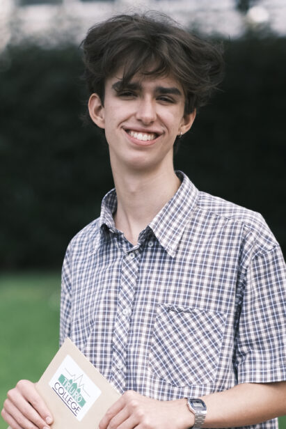 Student Gabe, smiles down camera, standing in a green area of the campus with a checked short sleeved shirt and his results envelope in hand