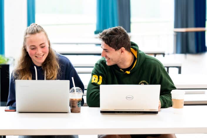 Two University students sit at a table with their laptops and cold drinks in the Fal building canteen.