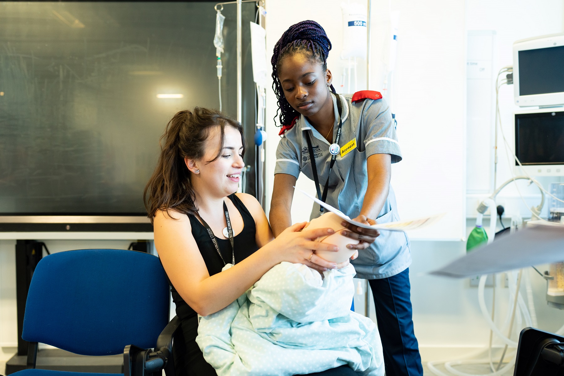 Two nursing students look at an infant training doll; one stands as the other sits.