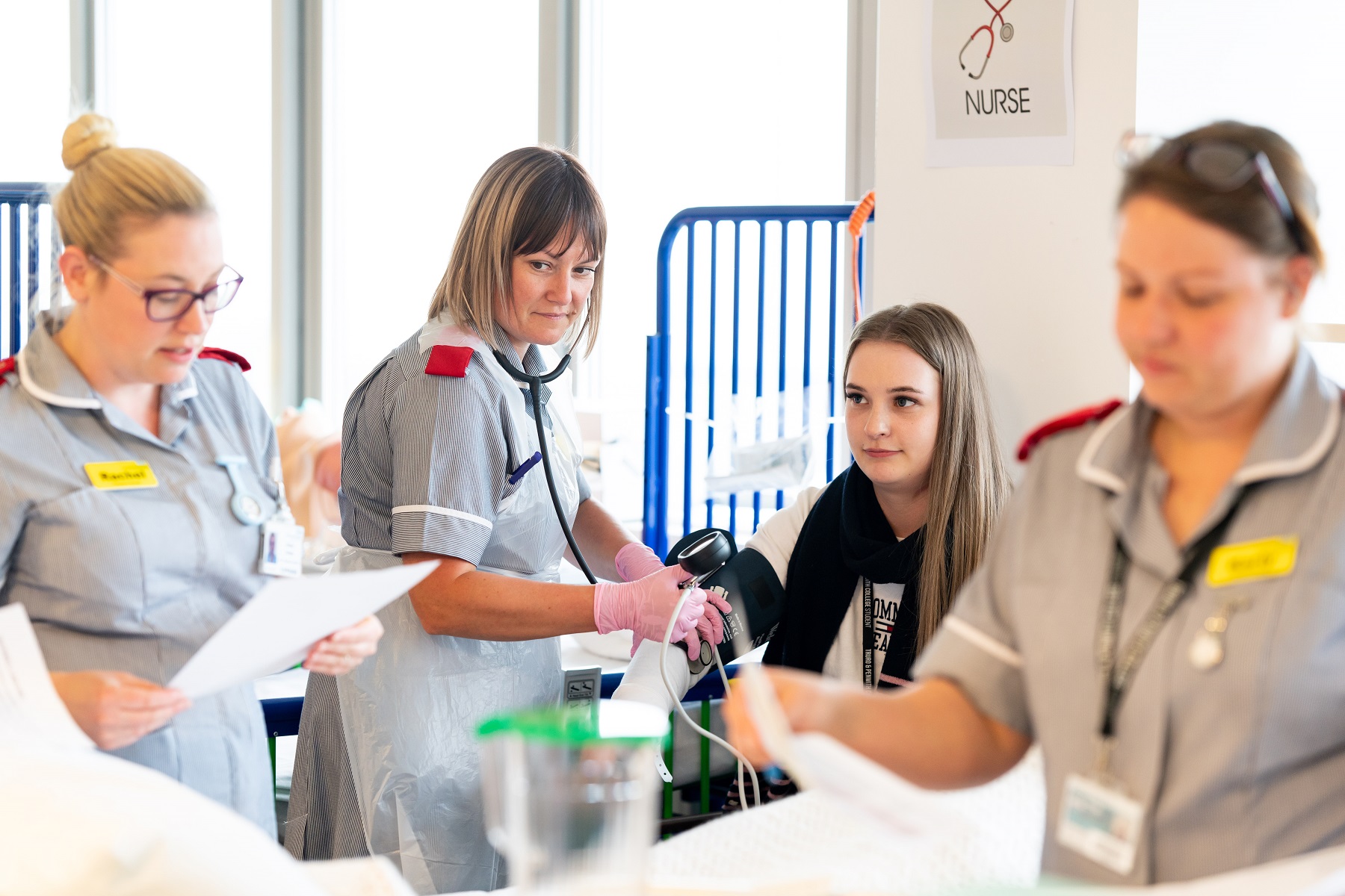 A nursing student takes the blood pressure of another student as two further students observe paperwork.