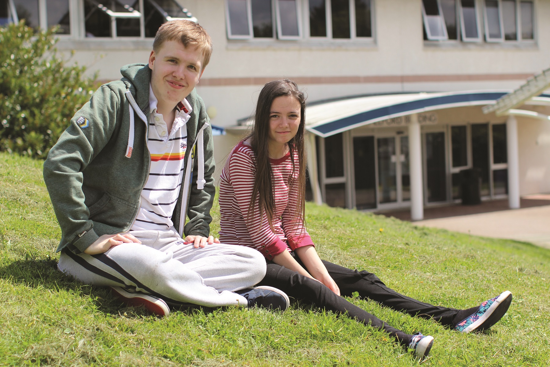 Two students sit on the grass outside the Helford building looking at the camera and smiling.