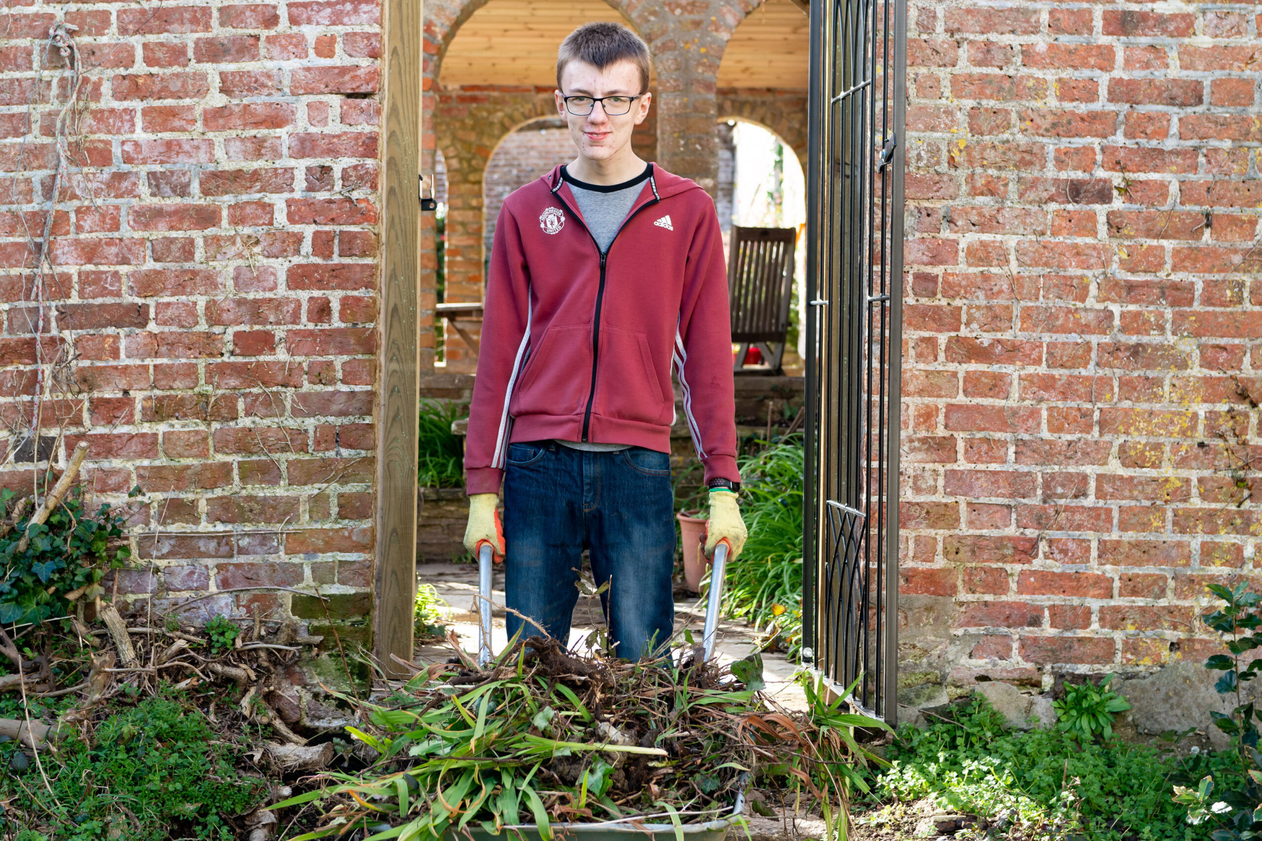 A student is pushing a wheelbarrow full of green waste through a doorway within a large garden wall.